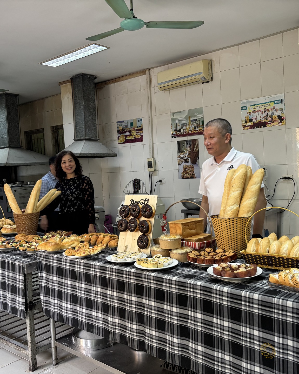 Quầy trưng bày bánh mì, bánh ngọt và donut do học viên Trường Hoa Sữa làm, hai khách tham quan đứng phía sau mỉm cười.
