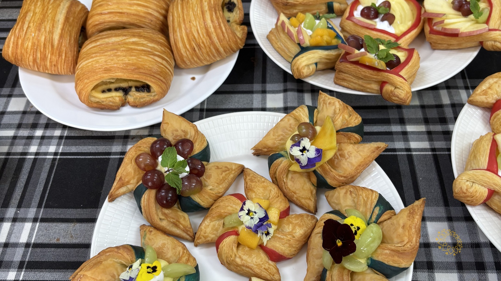 Cận cảnh các đĩa bánh sừng bò, bánh ngọt và bánh pastry trang trí nho, táo và hoa ăn được trên khăn trải bàn kẻ caro.