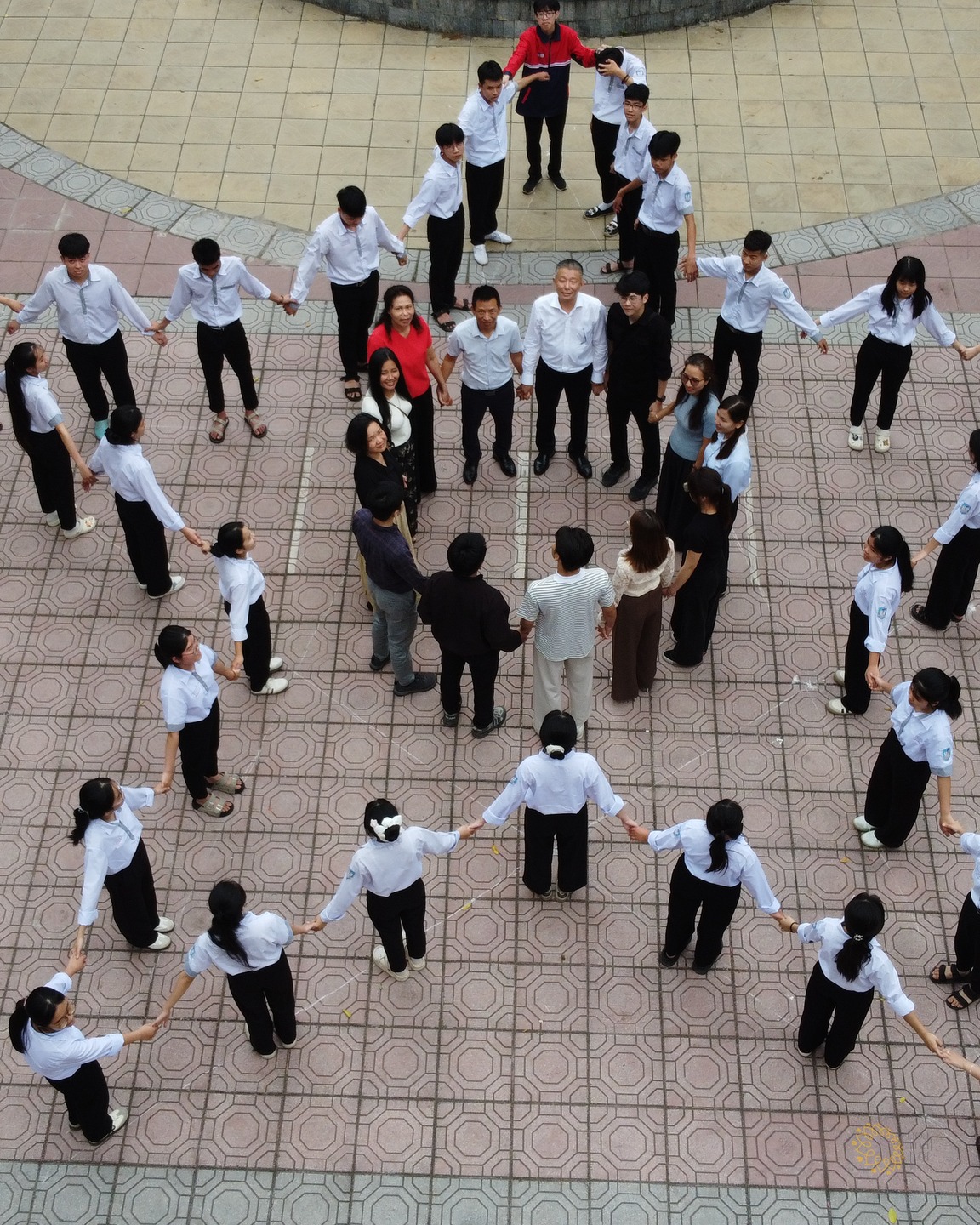 Toàn cảnh từ trên cao: các em học sinh cùng thầy cô tạo thành vòng tròn lớn nắm tay nhau trên sân gạch của trường.