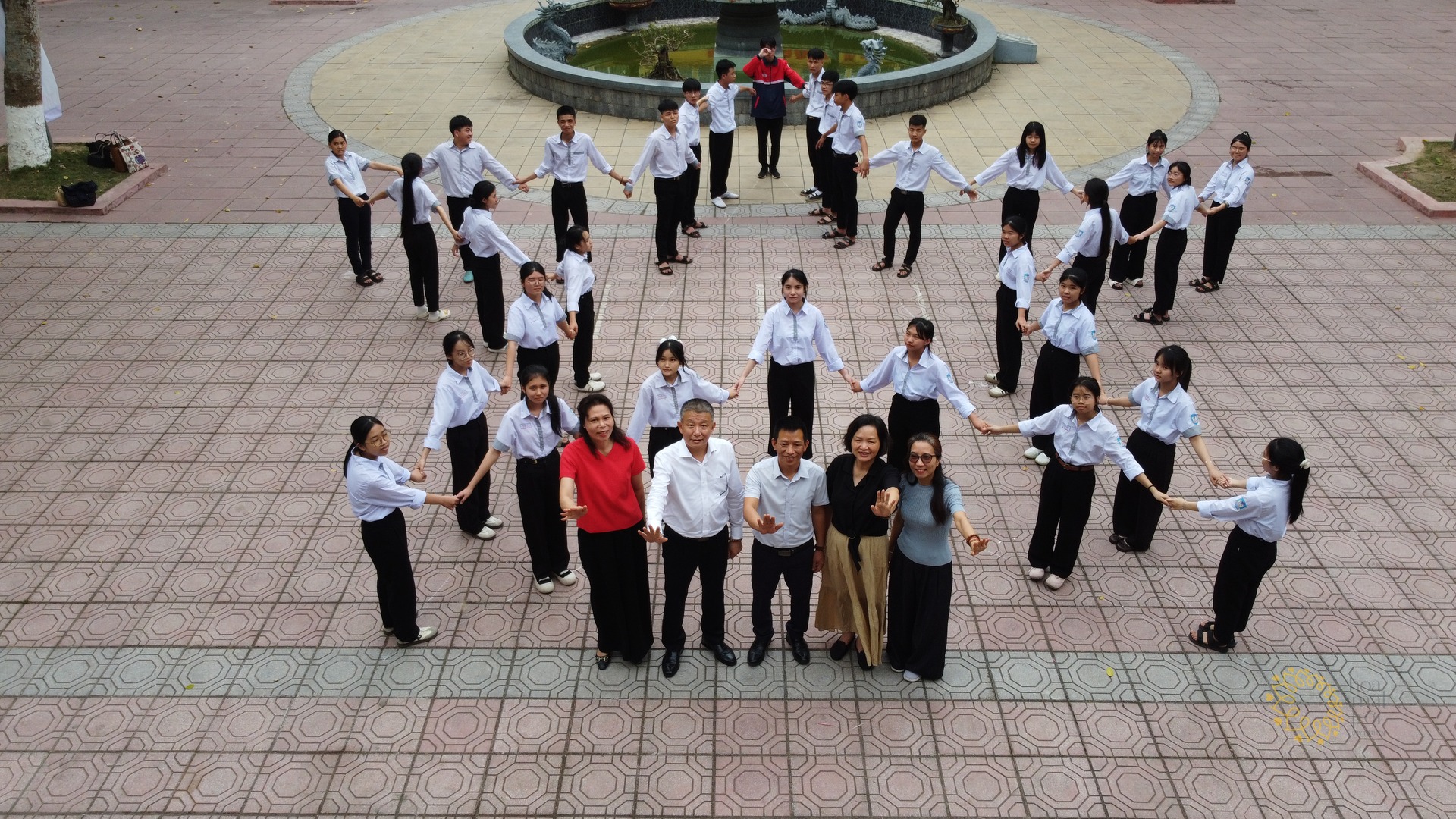 Nhìn từ trên cao: học sinh áo trắng và thầy cô đứng nắm tay nhau thành vòng tròn lớn quanh sân trường lát gạch.