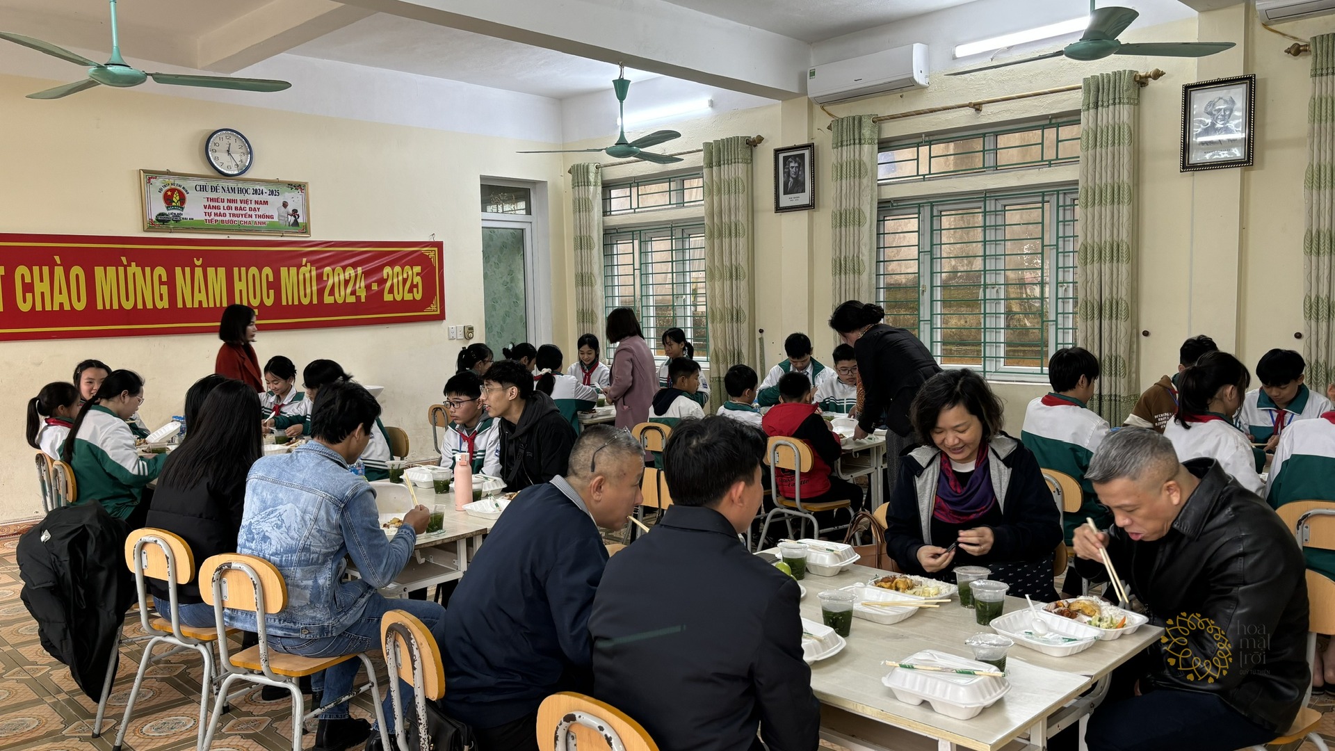 Học sinh THCS Đại An ngồi ăn trưa cùng thầy cô và thành viên Quỹ Hoa Mặt Trời trong phòng học có băng-rôn chào mừng năm học mới.