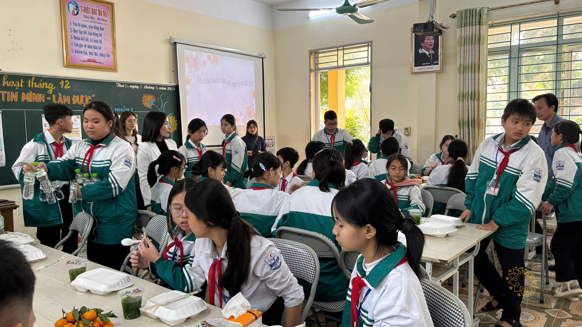 Các em học sinh mặc đồng phục xanh trắng quây quần bên những hộp cơm và đĩa quýt vàng, một bạn đứng chuyền chai nước cho bạn bên cạnh.