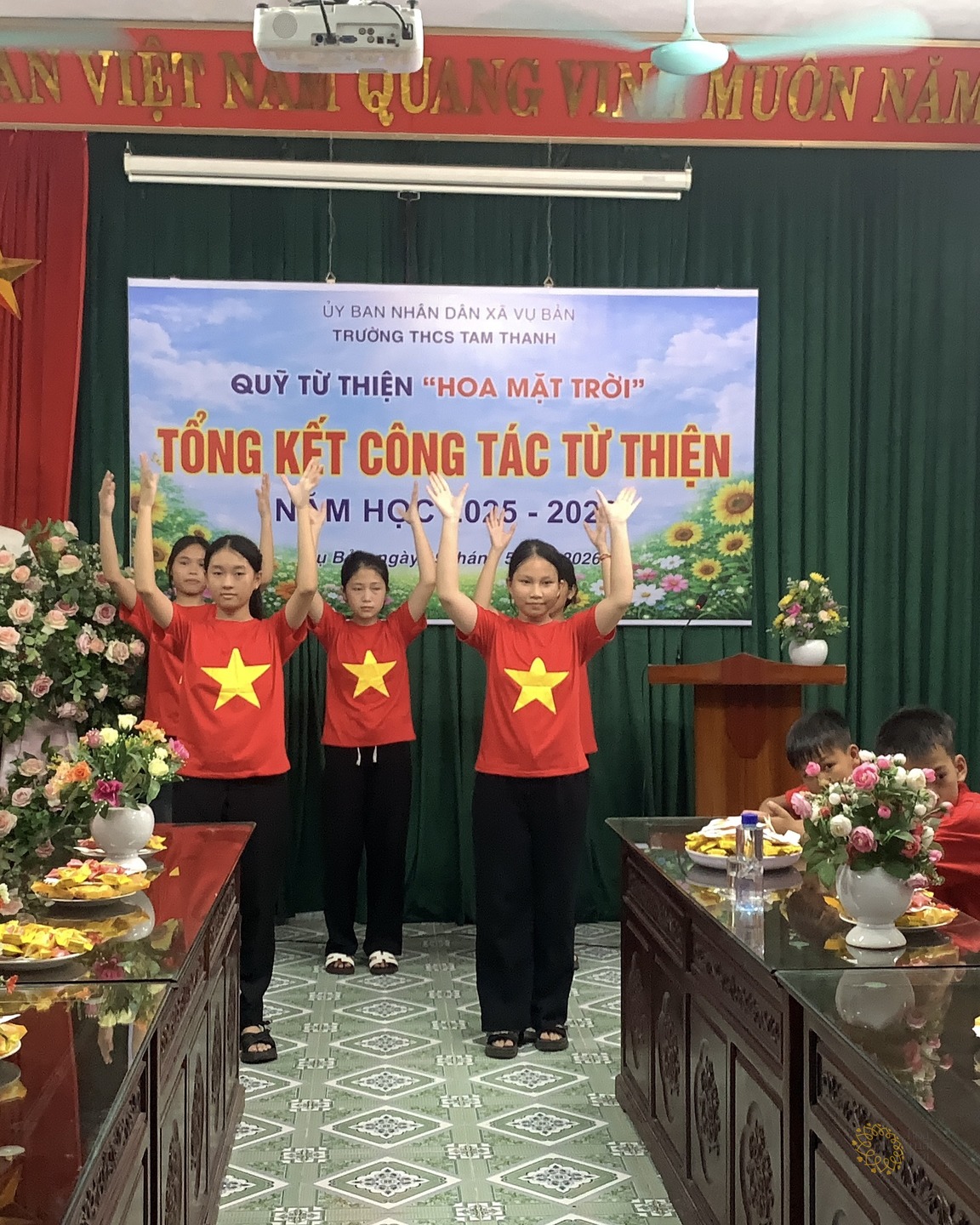 Ba em học sinh mặc áo cờ đỏ sao vàng đứng cạnh nhau trước phông "Tổng kết công tác từ thiện" của Quỹ Hoa Mặt Trời, cùng đưa hai tay lên trong một tiết mục văn nghệ; các bạn cùng lớp ngồi hai bên dãy bàn gỗ theo dõi.
