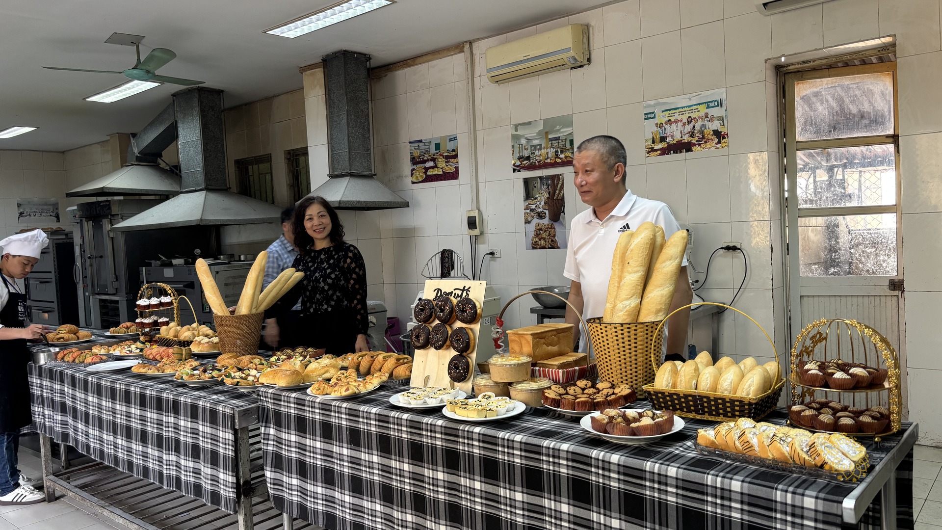 Quầy trưng bày bánh mì, bánh ngọt và donut do học viên Trường Hoa Sữa làm, hai khách tham quan đứng phía sau mỉm cười.