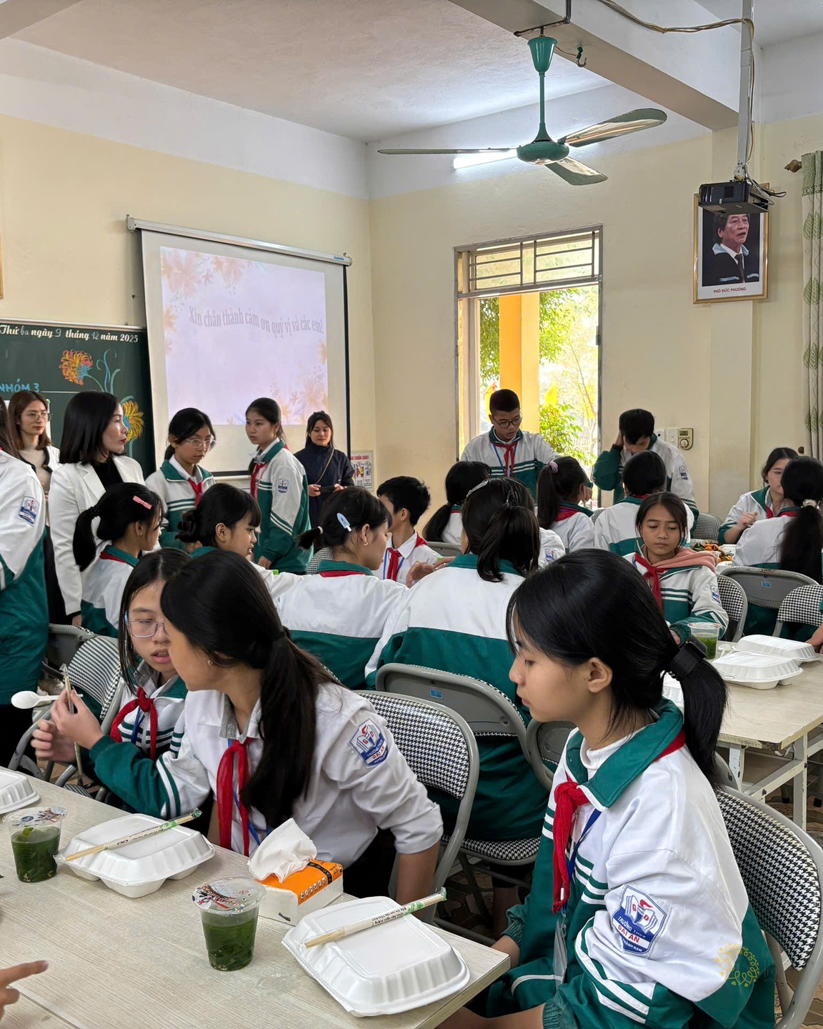 Các em học sinh mặc đồng phục xanh trắng quây quần bên những hộp cơm và đĩa quýt vàng, một bạn đứng chuyền chai nước cho bạn bên cạnh.