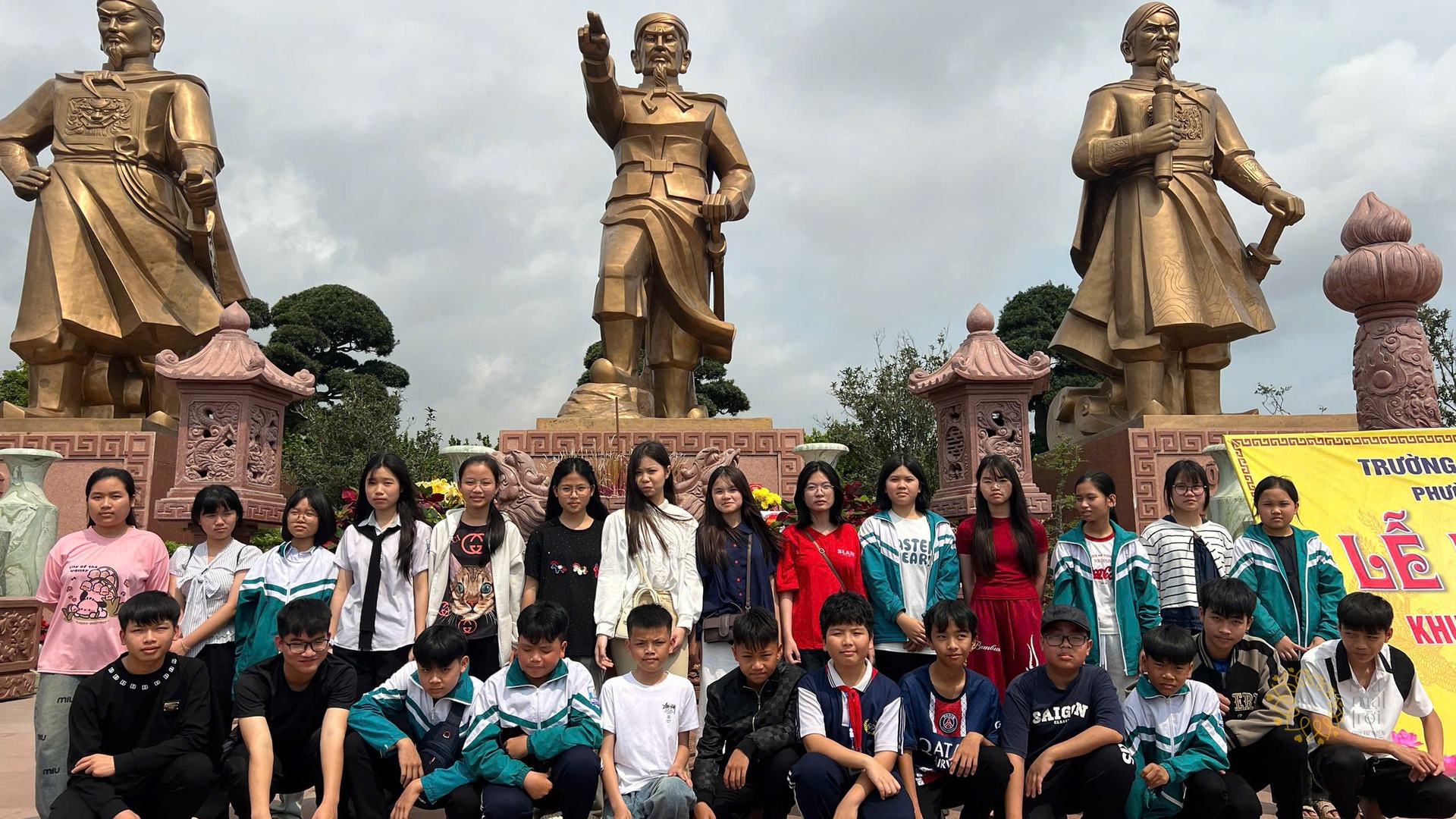Nhóm học sinh Tân Khánh đứng thành hai hàng trước cụm tượng đồng tại Bạch Đằng Giang, phía sau có băng-rôn vàng ghi "Lễ".