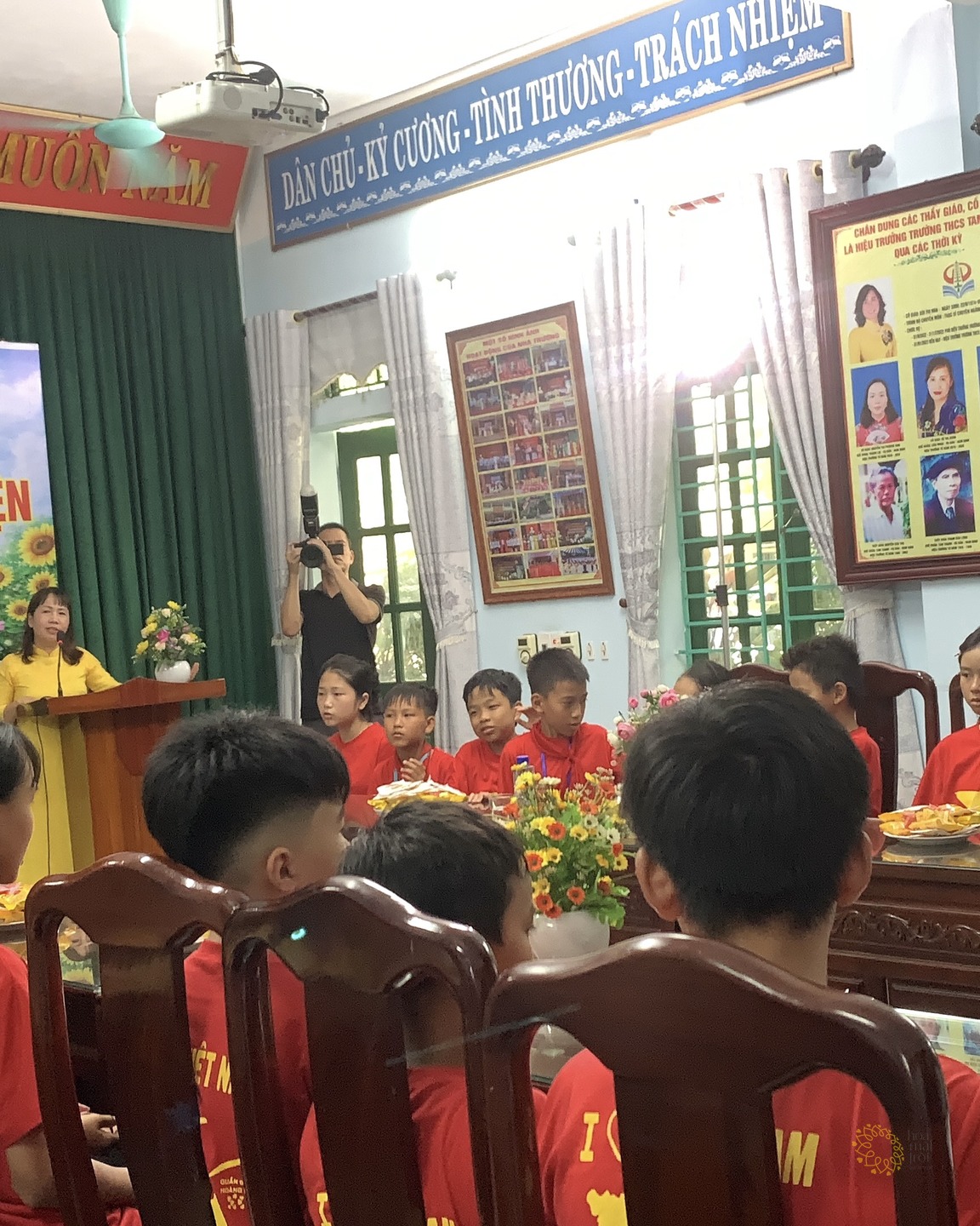 Một cô giáo mặc áo dài vàng đang phát biểu tại bục trong hội trường Trường THCS Tam Thanh; các em học sinh áo đỏ ngồi quây quanh các dãy bàn gỗ phía trước và bên phải.