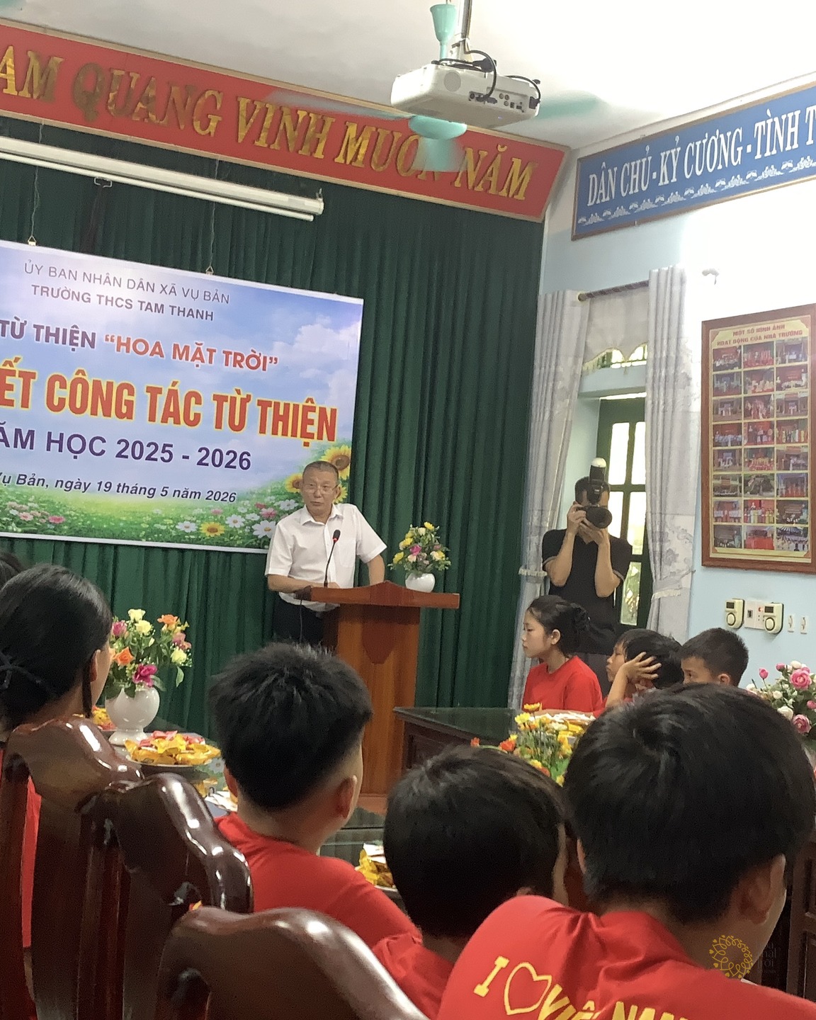 Một bác đại diện mặc áo sơ mi trắng đang phát biểu tại bục, phía sau là phông tổng kết ghi "Vụ Bản, ngày 19 tháng 5 năm 2026"; các em học sinh áo đỏ ngồi quanh các dãy bàn gỗ trong hội trường, một người đang cầm máy ảnh ghi hình ở bên phải.