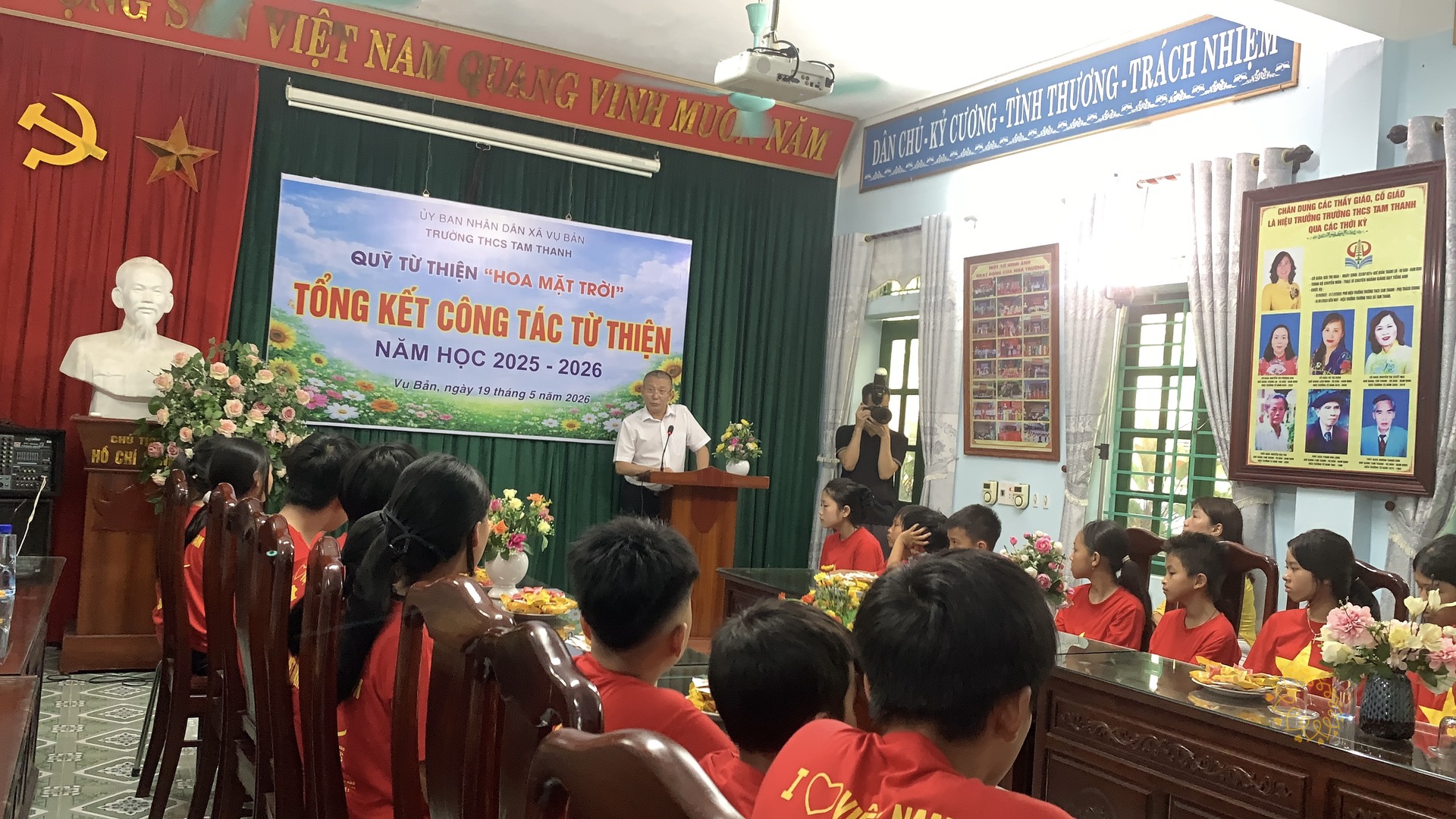 Một bác đại diện mặc áo sơ mi trắng đang phát biểu tại bục, phía sau là phông tổng kết ghi "Vụ Bản, ngày 19 tháng 5 năm 2026"; các em học sinh áo đỏ ngồi quanh các dãy bàn gỗ trong hội trường, một người đang cầm máy ảnh ghi hình ở bên phải.